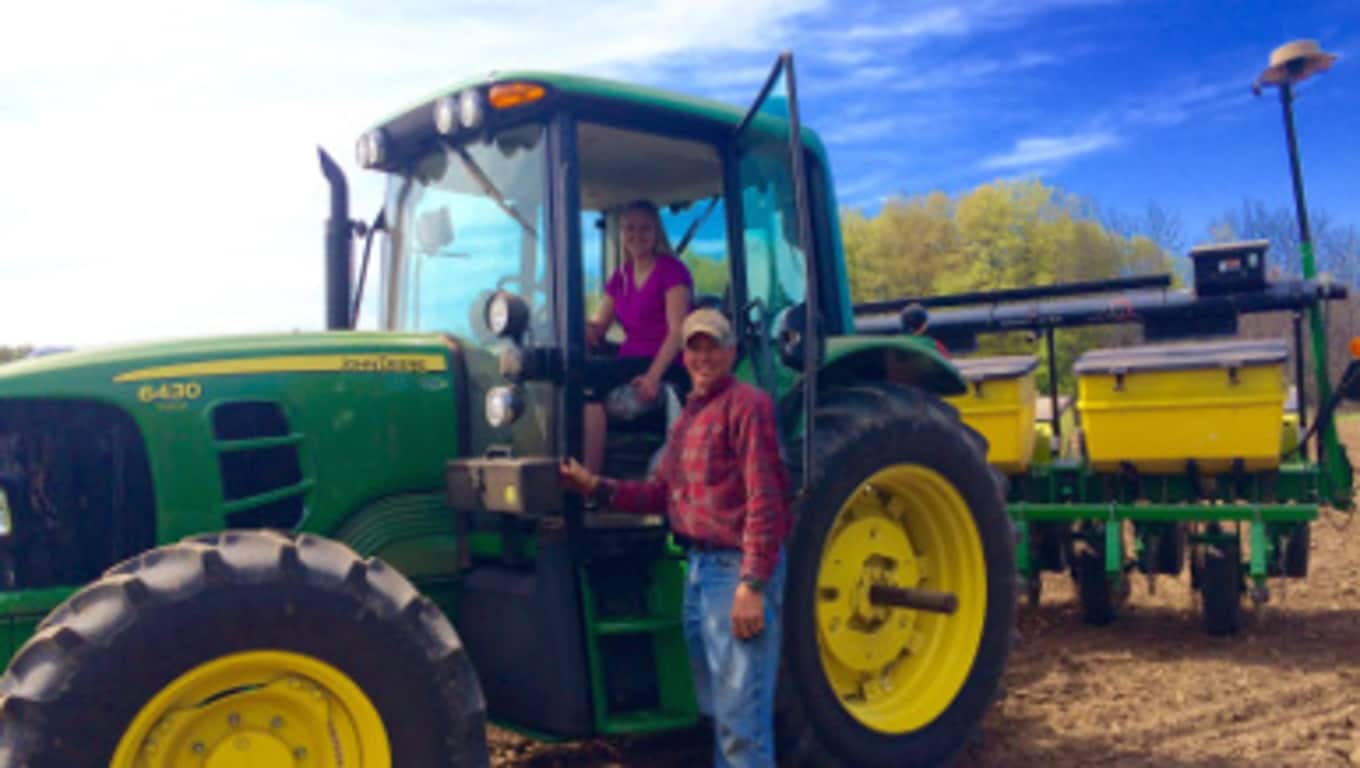 Hanna sits in a John Deere tractor, while her father stands