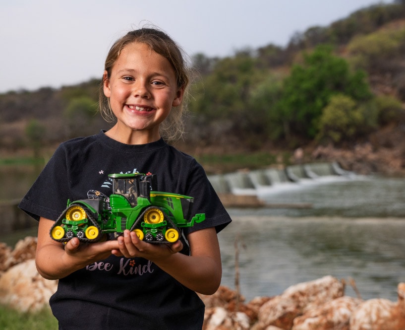 A child holds a green toy tractor while standing near a river with trees and rocks in the background.