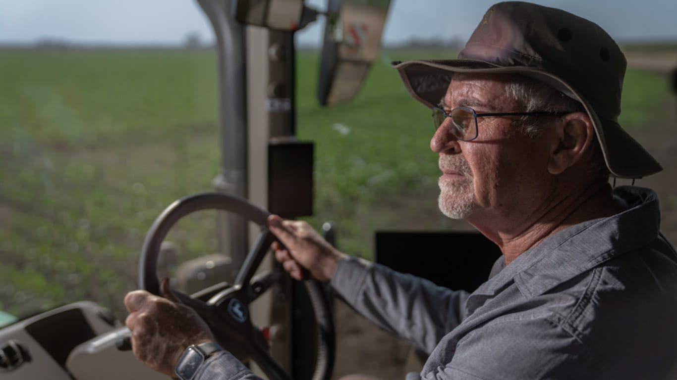Person operating a tractor from the cab, holding the steering wheel while driving beside a crop field.