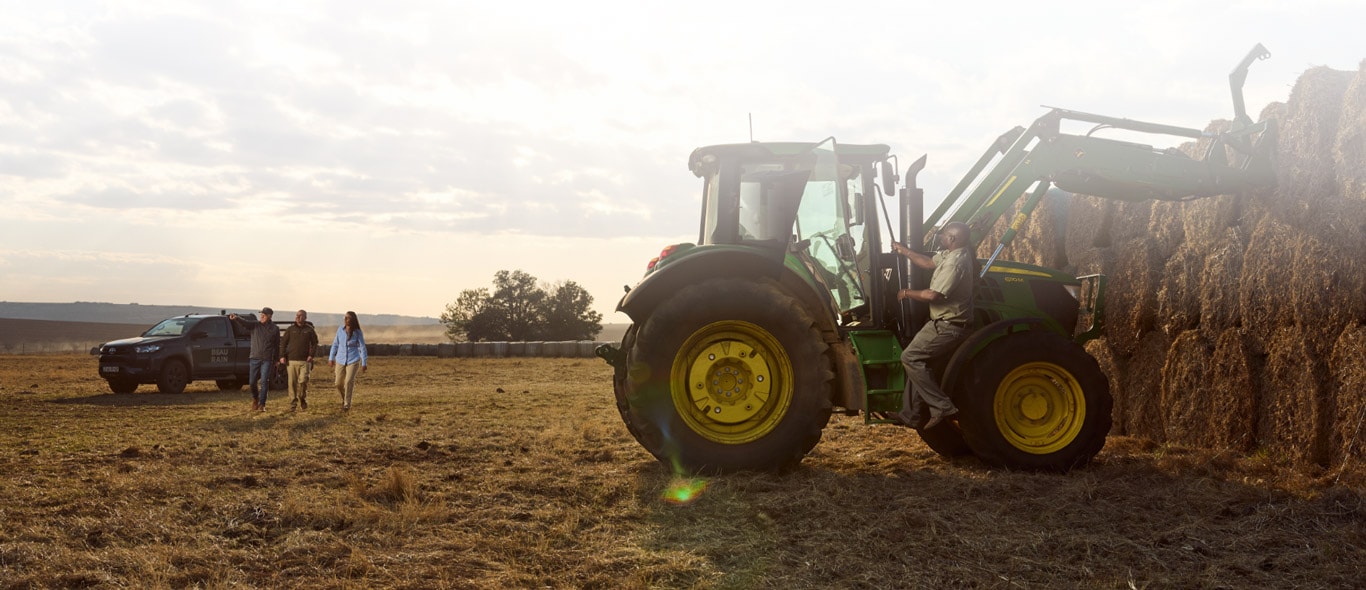A tractor lifts round hay bales while three people walk from a parked truck across an open field.