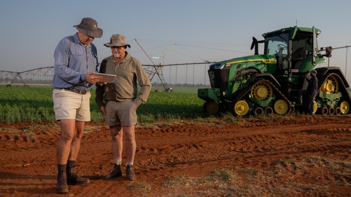 Two people review a tablet beside a tracked tractor near a crop field and a center pivot irrigation system.
